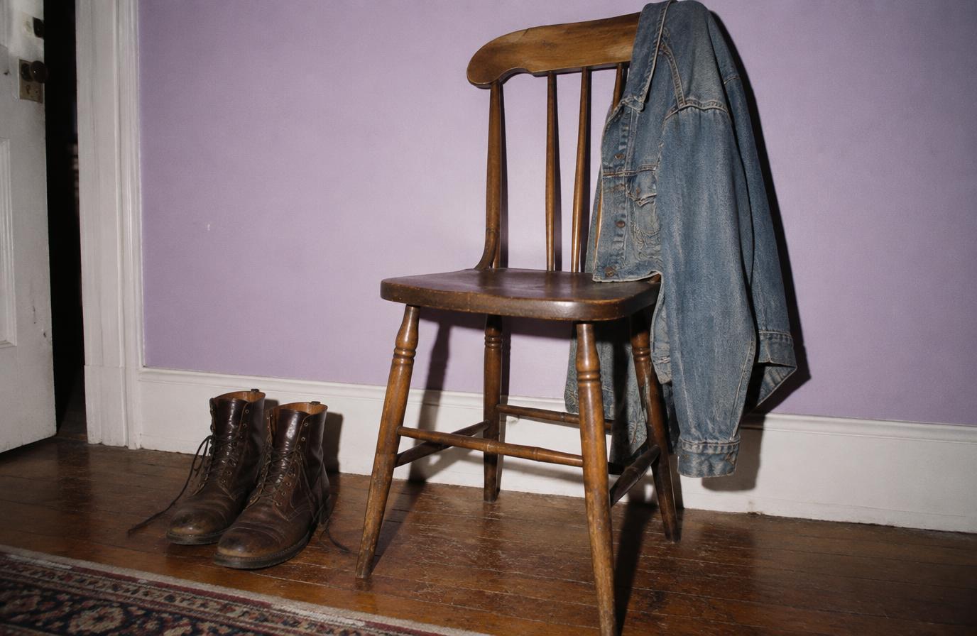 Wooden chair with a denim jacket over its back and leather boots beside it against a lilac wall